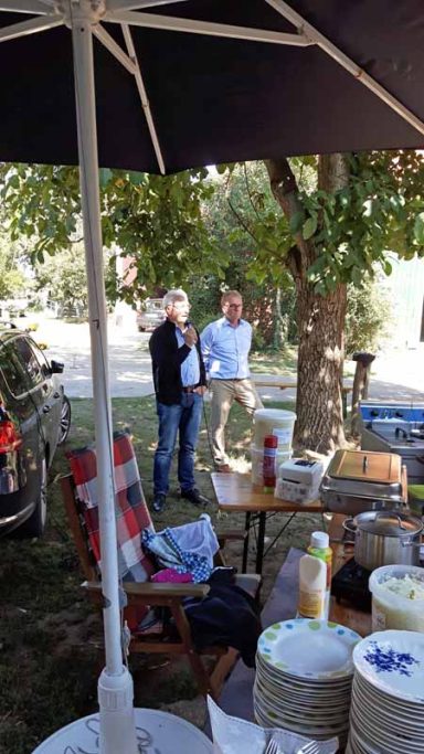Zwei Männer stehen unter einem Sonnenschirm in einem Garten. Tische mit Geschirr im Vordergrund.