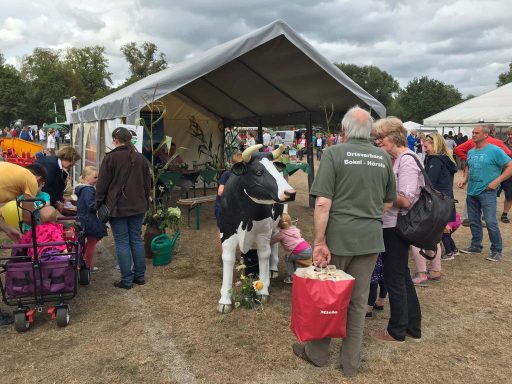 Menschen auf einem Fest mit Ständen, einer Kuhfigur und bunten Blumen.