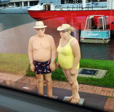 Zwei lebensgroße, figurative Skulpturen eines Paares in Badebekleidung stehen am Hafen.