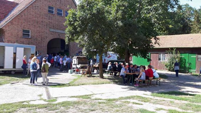 Menschen versammeln sich im Freien vor einem Gebäude, unter einem Baum in einer ländlichen Umgebung.