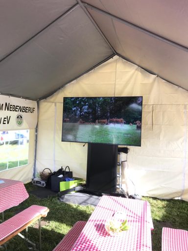 Ein Fernseher zeigt ein Bild in einem Zelt mit Bänken und Tisch.