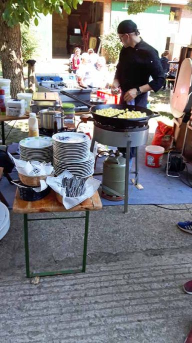 Ein Koch bereitet Essen an einem Grill im Freien vor, umgeben von Tischen und Geräten.